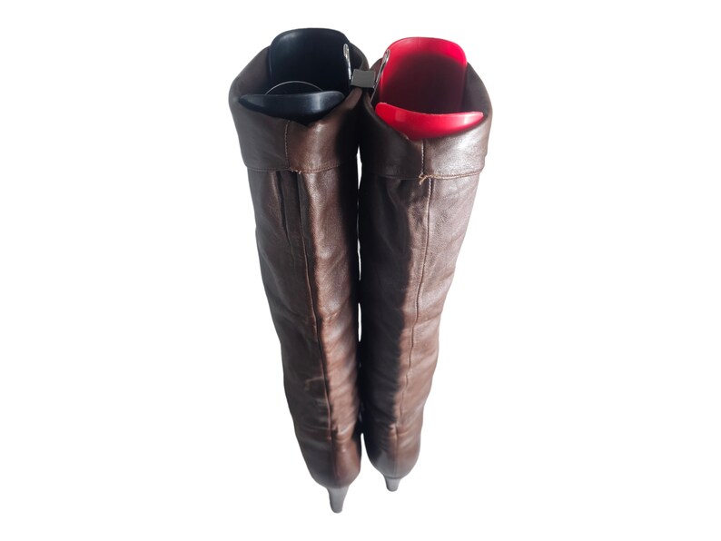 May include: A pair of brown leather knee-high boots with a pointed toe. The boots are folded down at the top, revealing a red and black shoe stretcher inside.