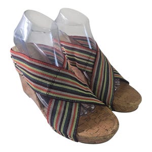 Puede incluir: Par de sandalias de cuña con suela de corcho y correas a rayas multicolores. Las correas se cruzan sobre el pie. El tacón de cuña mide aproximadamente 7.6 cm de alto.