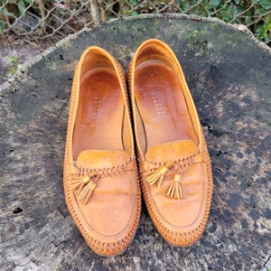 May include: A pair of brown leather tassel loafers with a woven detail around the edge. The loafers are on a wooden surface.