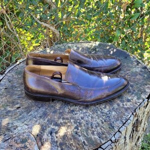 Pode incluir: Um par de sapatos marrons de couro com bico fino. Os sapatos est&atilde;o sobre uma superf&iacute;cie de madeira.