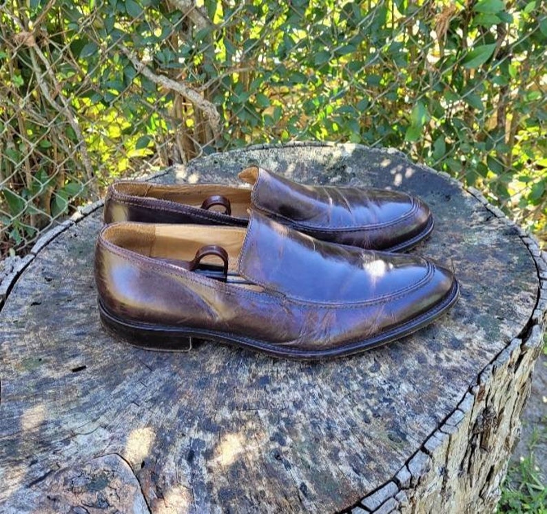Pode incluir: Um par de sapatos marrons de couro com bico fino. Os sapatos est&atilde;o sobre uma superf&iacute;cie de madeira.