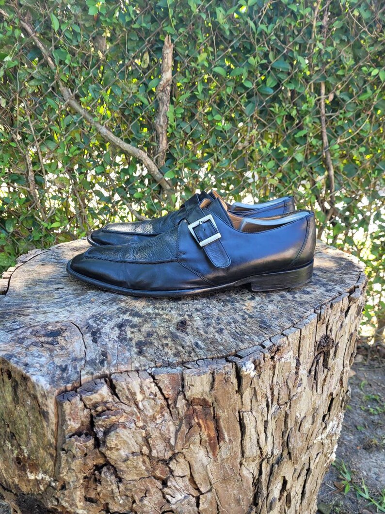K&ouml;nnte beinhalten: Ein Paar schwarze Lederschuhe mit einem einzigen Schnallenriemen. Die Schuhe stehen auf einem Baumstumpf.