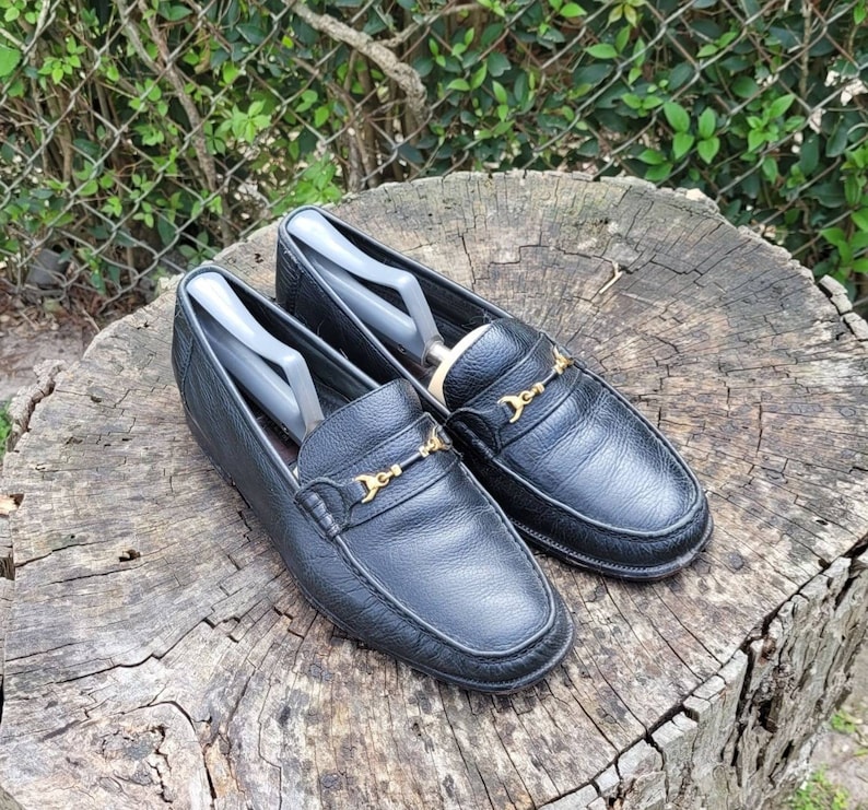 May include: A pair of black leather loafers with gold buckles. The loafers are on a wooden surface.
