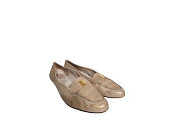 Puede incluir: Un par de mocasines de cuero beige con empeine texturizado y un acento rectangular dorado. Los zapatos tienen un diseño clásico sin cordones y están sobre un fondo blanco.