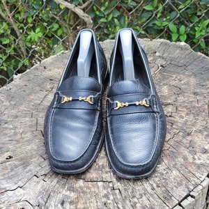 May include: A pair of black leather loafers with gold metal buckles. The loafers are on a wooden surface.