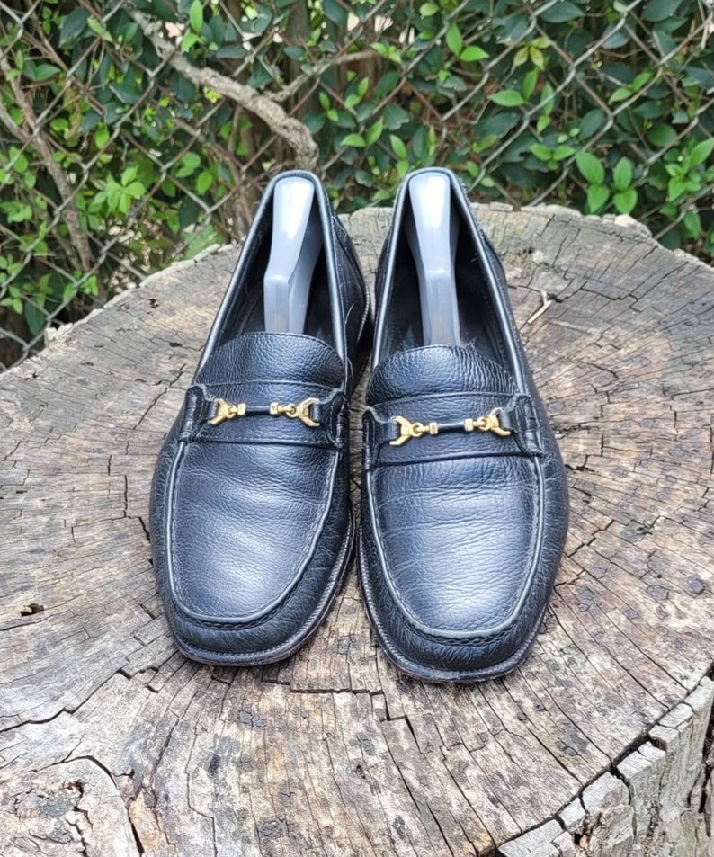 May include: A pair of black leather loafers with gold metal buckles. The loafers are on a wooden surface.