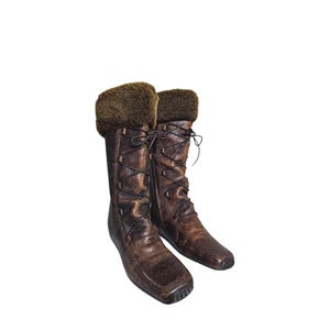 Puede incluir: Un par de botas de cuero marrón con un acabado texturizado. Las botas tienen un diseño con cordones y un ribete de piel sintética en la parte superior. Las botas son hasta la rodilla y tienen punta cuadrada.
