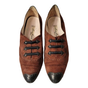 May include: A pair of brown suede shoes with black trim and laces. The shoes have a pointed toe and a black rubber sole. The shoes are labeled "Miss Maud Paris" and "Made in Italy".