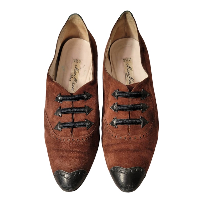 May include: A pair of brown suede shoes with black trim and laces. The shoes have a pointed toe and a black rubber sole. The shoes are labeled "Miss Maud Paris" and "Made in Italy".