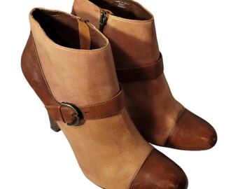 May include: A pair of brown leather ankle boots with a buckle detail. The boots have a light brown upper and a darker brown toe cap. The boots are made in Italy.