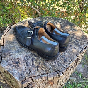K&ouml;nnte beinhalten: Ein Paar schwarze Monk-Strap-Schuhe aus Leder mit einer silbernen Schnalle. Die Schuhe stehen auf einem Baumstumpf.