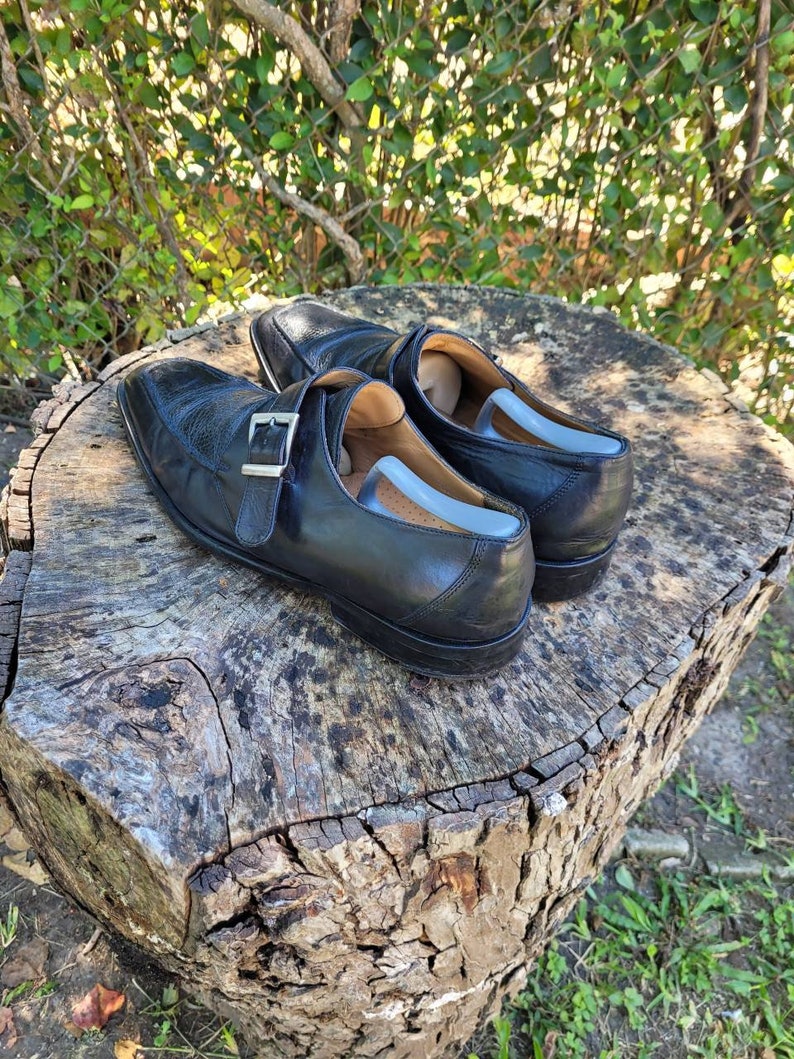 K&ouml;nnte beinhalten: Ein Paar schwarze Monk-Strap-Schuhe aus Leder mit einer silbernen Schnalle. Die Schuhe stehen auf einem Baumstumpf.