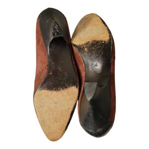 May include: A pair of brown suede heeled shoes with black soles. The shoes have a pointed toe and a small heel.