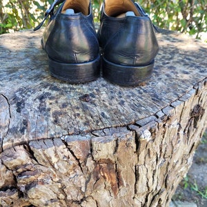 K&ouml;nnte beinhalten: Ein Paar schwarze Monk-Strap-Schuhe aus Leder mit silbernen Schnallen. Die Schuhe stehen auf einem Baumstumpf.