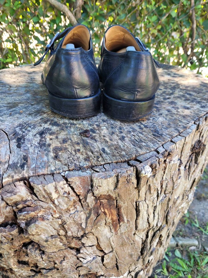 K&ouml;nnte beinhalten: Ein Paar schwarze Monk-Strap-Schuhe aus Leder mit silbernen Schnallen. Die Schuhe stehen auf einem Baumstumpf.