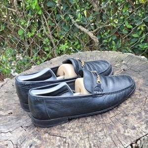 May include: A pair of black leather loafers with gold buckles. The loafers are on a wooden surface.