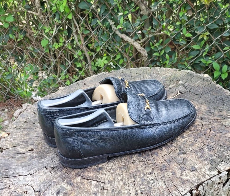 May include: A pair of black leather loafers with gold buckles. The loafers are on a wooden surface.