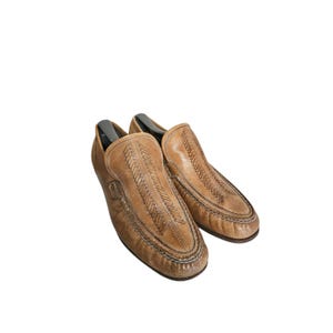 May include: A pair of tan leather loafers featuring a moccasin-style construction with decorative stitched detailing along the vamp and a rounded toe. The shoes include black plastic shoe trees inserted inside to maintain their shape.