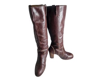 May include: A pair of brown leather knee-high boots with a block heel. The boots have a white stitching detail around the top and heel.