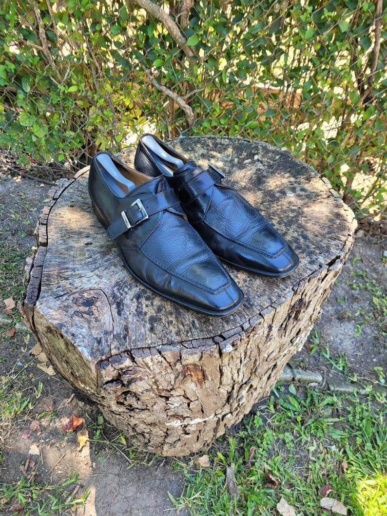 K&ouml;nnte beinhalten: Ein Paar schwarze Monk-Strap-Schuhe aus Leder mit Schnallenverschluss. Die Schuhe stehen auf einem Baumstumpf.