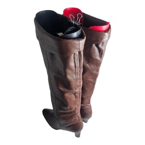 May include: A pair of brown leather knee-high boots with a heel. The boots are being held up by a red boot shaper.
