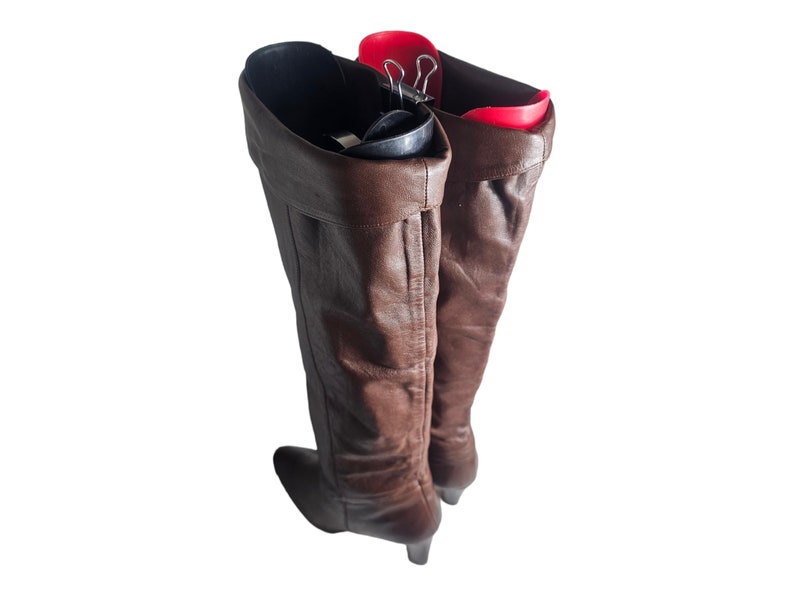 May include: A pair of brown leather knee-high boots with a heel. The boots are being held up by a red boot shaper.