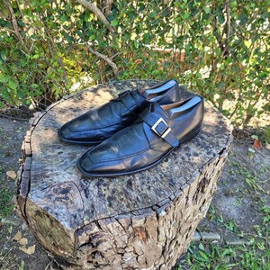 K&ouml;nnte beinhalten: Ein Paar schwarze Lederschuhe mit Schnallenverschluss. Die Schuhe stehen auf einem Baumstumpf.