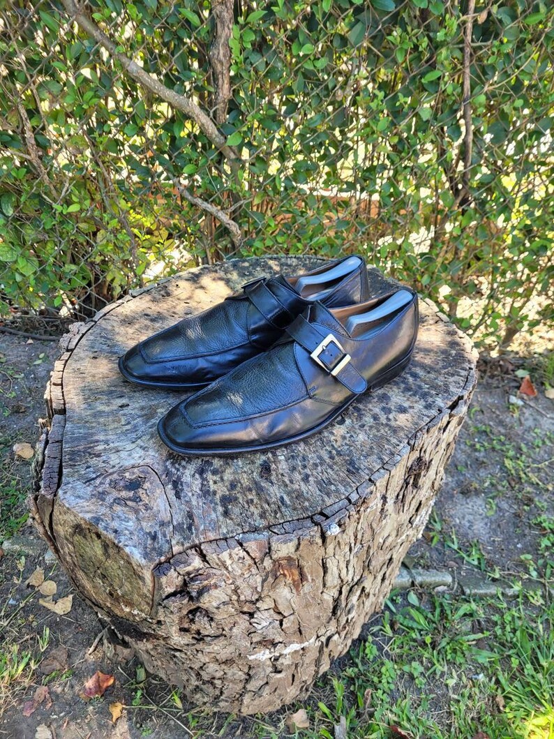 K&ouml;nnte beinhalten: Ein Paar schwarze Lederschuhe mit Schnallenverschluss. Die Schuhe stehen auf einem Baumstumpf.