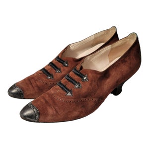 May include: Brown suede heeled shoes with black ribbon laces and decorative metal accents. The shoes have a pointed toe and a low heel.