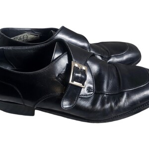 Pode incluir: Um par de sapatos de monge de couro preto com fivela prateada. Os sapatos t&ecirc;m bico fino e salto baixo.