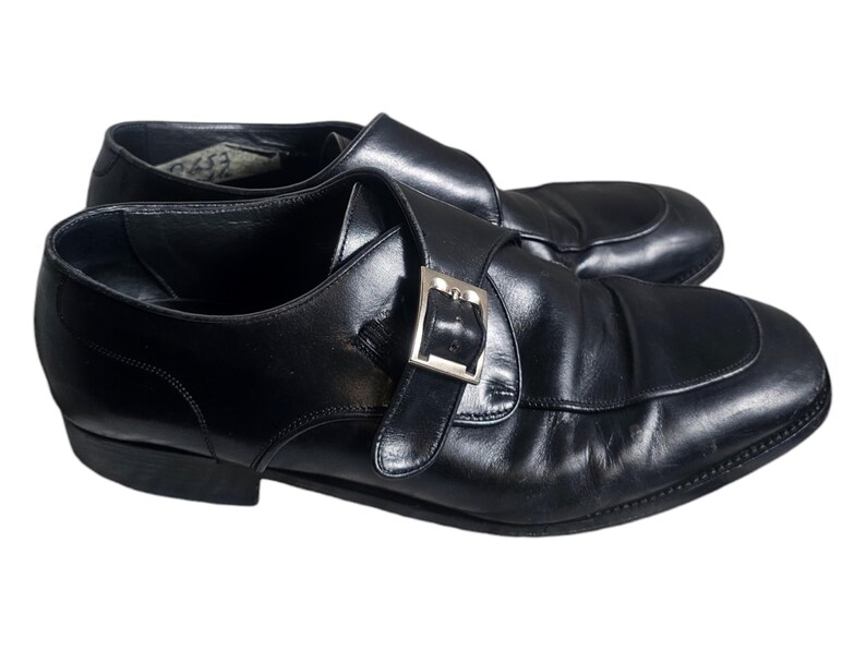 Pode incluir: Um par de sapatos de monge de couro preto com fivela prateada. Os sapatos t&ecirc;m bico fino e salto baixo.