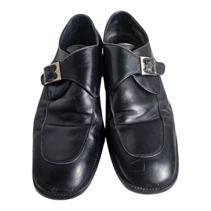 Pode incluir: Um par de sapatos de couro preto com uma &uacute;nica fivela. Os sapatos t&ecirc;m bico fino e salto baixo.