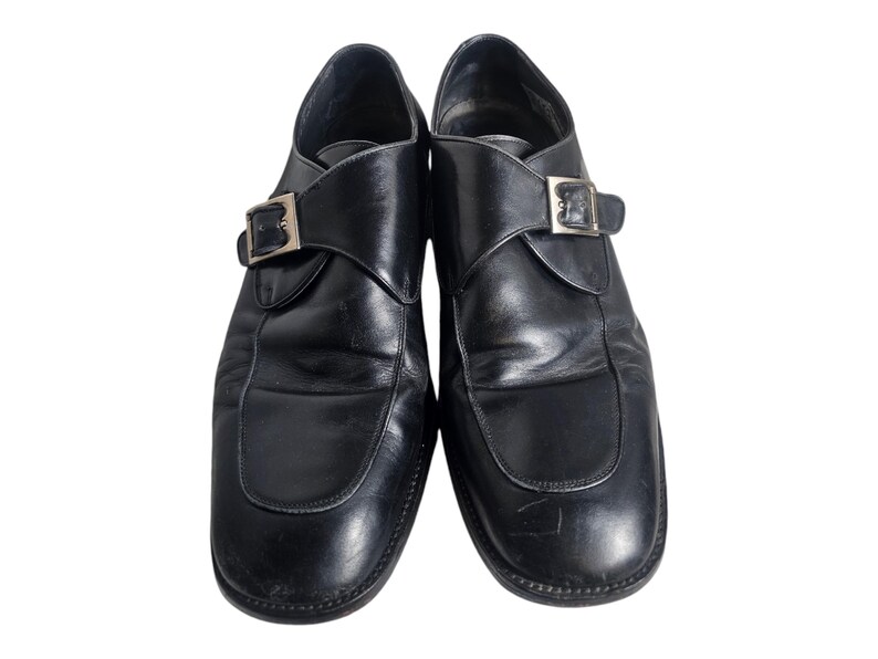 Pode incluir: Um par de sapatos de couro preto com uma &uacute;nica fivela. Os sapatos t&ecirc;m bico fino e salto baixo.