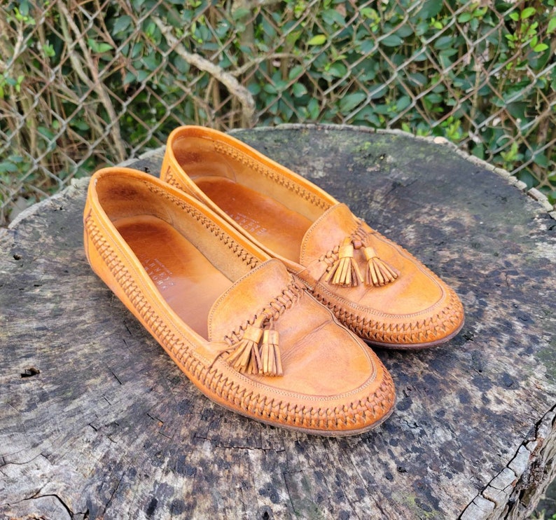 May include: A pair of brown leather tassel loafers with a woven stitch detail. The loafers have a pointed toe and a tassel on each shoe.