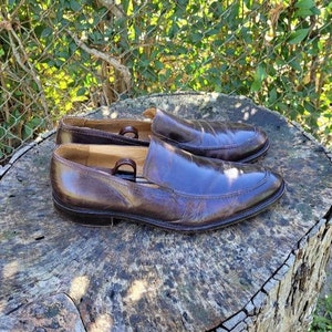 Pode incluir: Um par de sapatos marrons de couro com bico fino. Os sapatos est&atilde;o sobre uma superf&iacute;cie de madeira.