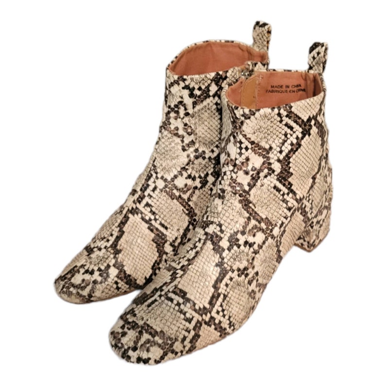 May include: A pair of ankle boots with a snakeskin pattern. The boots are beige and brown with a block heel. The label on the boot reads "Made in China, Fabrique en Chine."