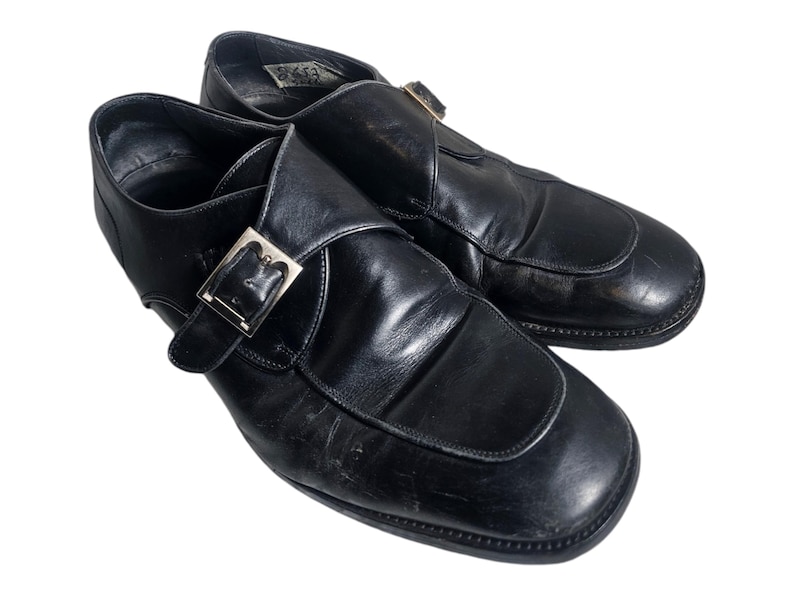Pode incluir: Um par de sapatos de couro preto com fecho de fivela. Os sapatos t&ecirc;m bico fino e salto baixo.