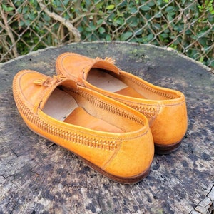 May include: A pair of brown leather tassel loafers with a woven detail around the edge. The loafers are sitting on a wooden surface.