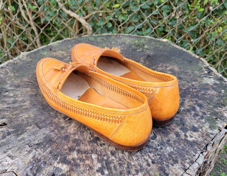 May include: A pair of brown leather tassel loafers with a woven detail around the edge. The loafers are sitting on a wooden surface.