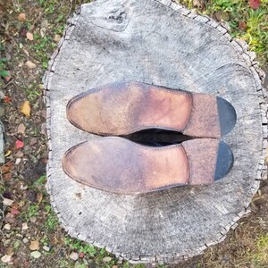 Puede incluir: Un par de zapatos de cuero marr&oacute;n con suelas negras. Los zapatos est&aacute;n colocados sobre un toc&oacute;n de &aacute;rbol con una corteza &aacute;spera y gris.