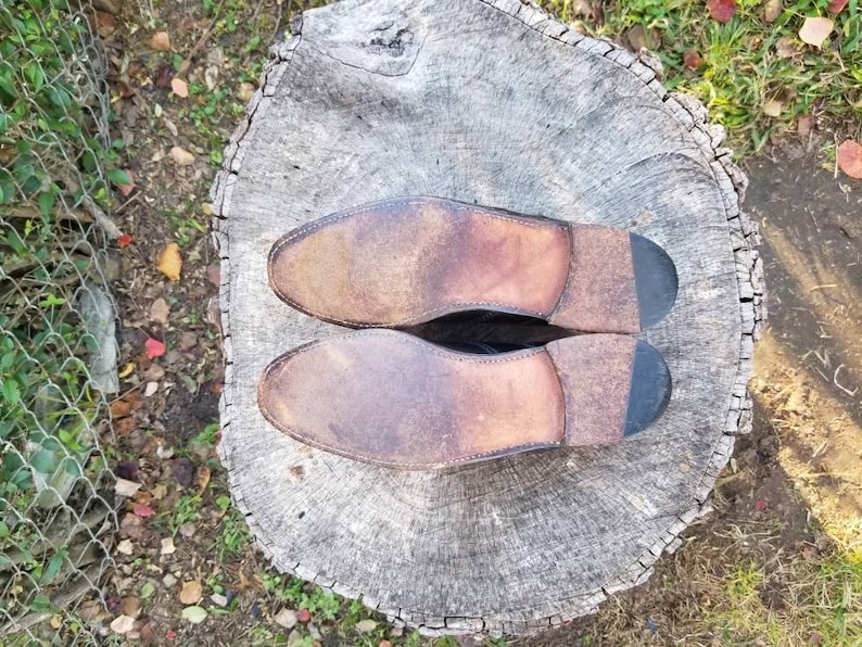 Puede incluir: Un par de zapatos de cuero marr&oacute;n con suelas negras. Los zapatos est&aacute;n colocados sobre un toc&oacute;n de &aacute;rbol con una corteza &aacute;spera y gris.