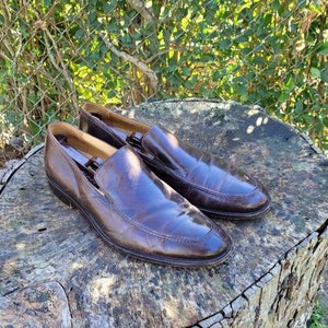 Pode incluir: Um par de sapatos marrons de couro com bico fino. Os sapatos est&atilde;o sobre uma superf&iacute;cie de madeira.