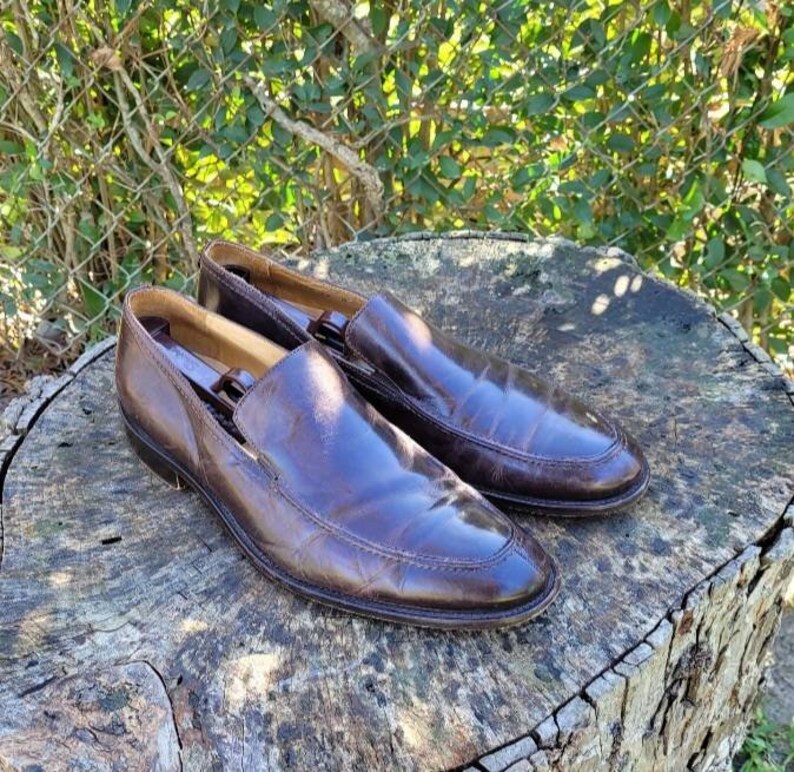 Pode incluir: Um par de sapatos marrons de couro com bico fino. Os sapatos est&atilde;o sobre uma superf&iacute;cie de madeira.