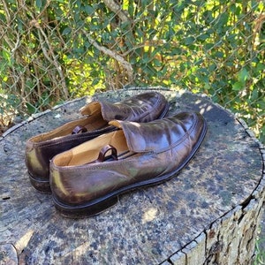 Pode incluir: Um par de mocassins de couro marrom com um design de deslizamento. Os mocassins est&atilde;o sentados em um toco de &aacute;rvore.
