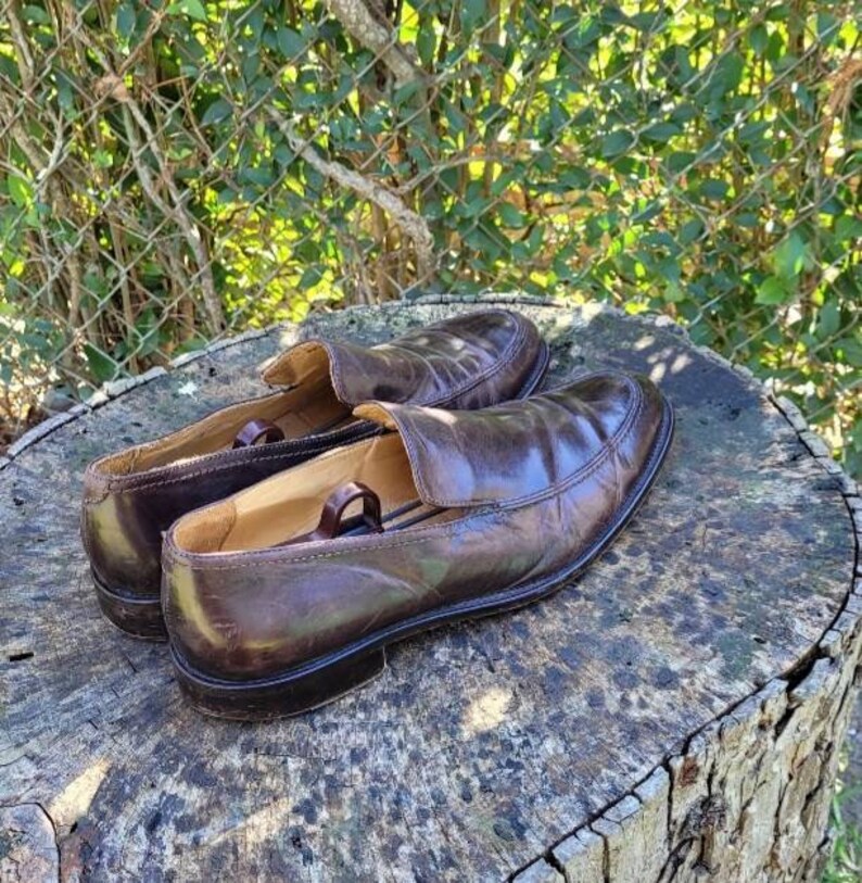 Pode incluir: Um par de mocassins de couro marrom com um design de deslizamento. Os mocassins est&atilde;o sentados em um toco de &aacute;rvore.