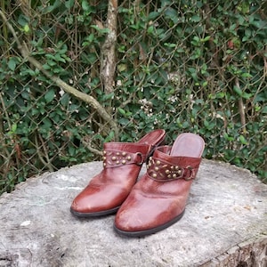 Op de afbeelding: Een paar bruine leren muiltjes met gouden gesp met studs. De schoenen staan op een boomstronk.
