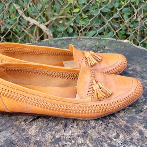 May include: A pair of brown leather tassel loafers with a woven pattern on the upper. The loafers are sitting on a wooden surface.