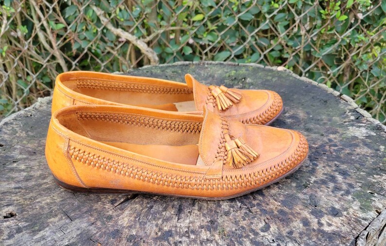 May include: A pair of brown leather tassel loafers with a woven pattern on the upper. The loafers are sitting on a wooden surface.
