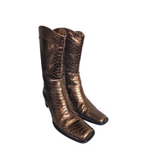 Puede incluir: Un par de botas hasta la rodilla con un estampado de piel de serpiente bronce metálico. Las botas tienen punta cuadrada y tacón pequeño. Las botas están hechas de un material brillante y parecen ser de cuero.