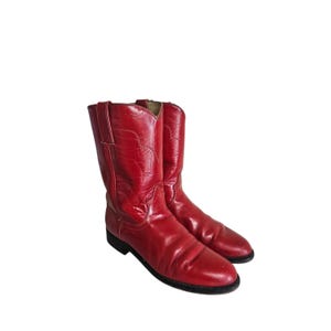 Könnte beinhalten: Ein Paar wadenhohe Stiefel in leuchtendem Rot mit glatter, polierter Oberfläche. Die Stiefel verfügen über eine abgerundete Zehenpartie, einen niedrigen Blockabsatz und dekorative Ziernähte entlang des Schafts und Fußes sowie eine schwarze Sohle.